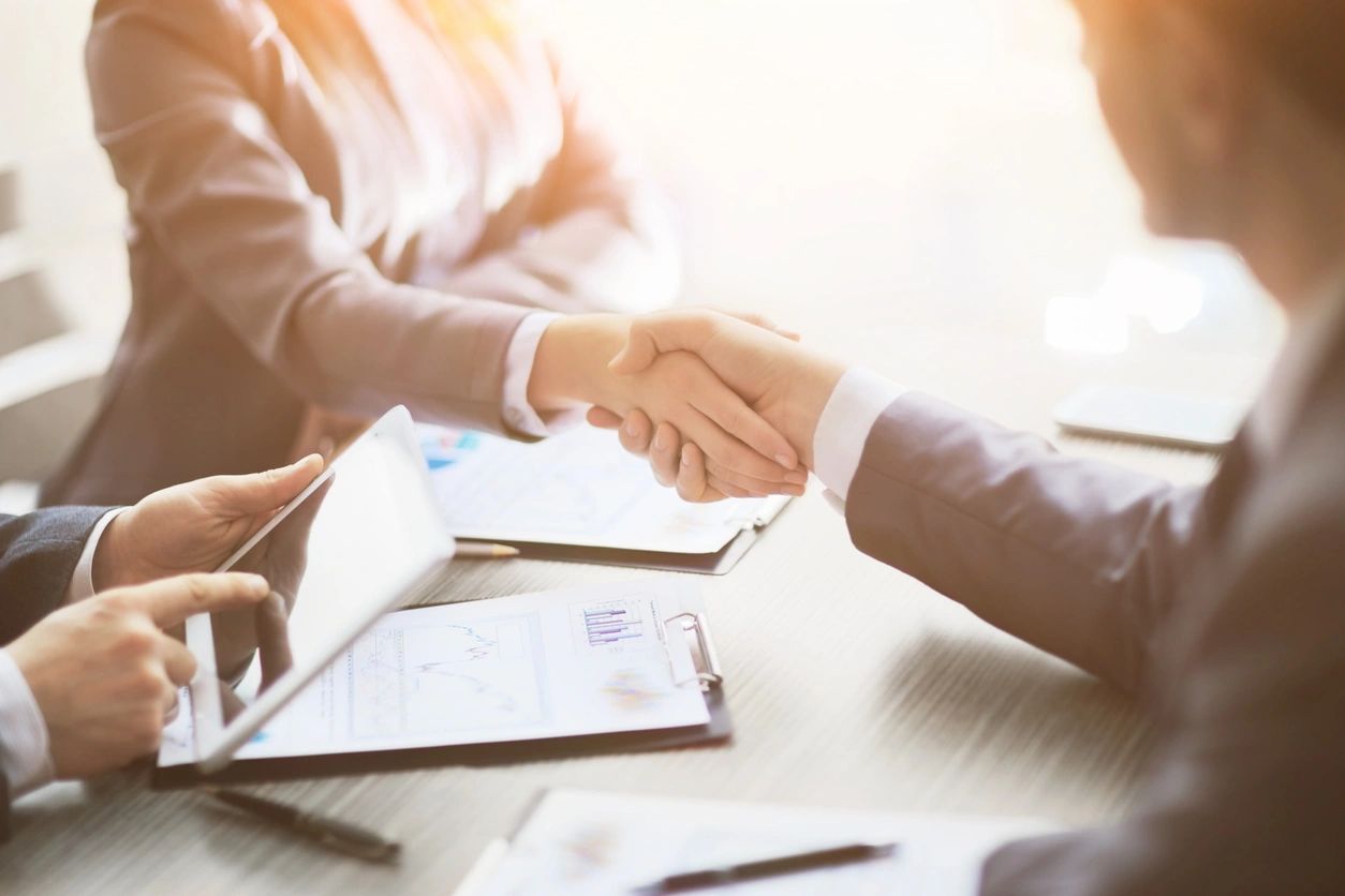 A businessman and business woman shaking hands in front of another businessman using a tablet.