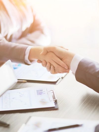 Two people shaking hands over a table of documents with the sun shining on them. 