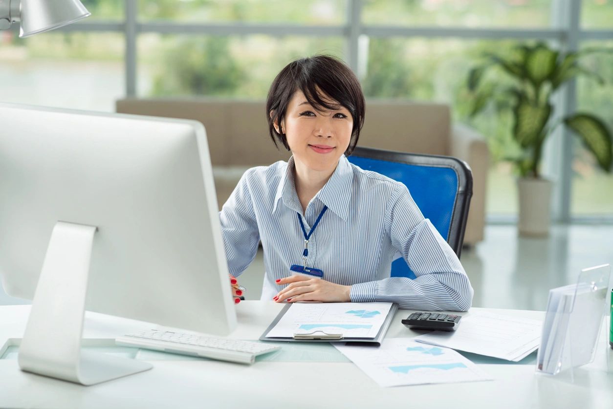 A lady presents a welcoming smile as she sits at her desk with her clipboard, calculator, keyboard and computer monitor in front of her.