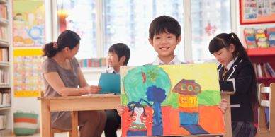 A smiling boy proudly shows his colorful drawing in a classroom.