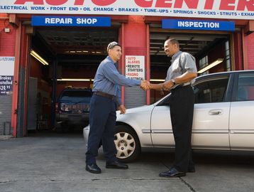 Two men shaking hands outside a car repair shop beside a white car.