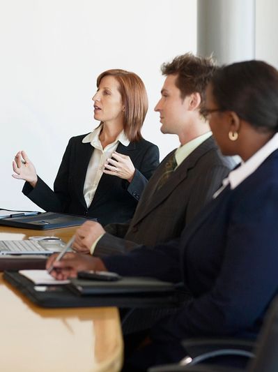 Two women and a man, all in suits, at a conference table in a meeting, gesturing and writing.