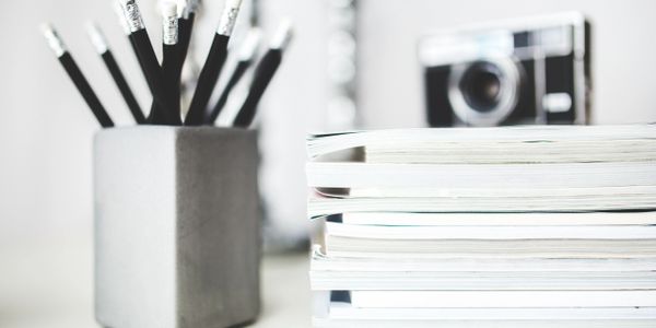 Pencils in a pot next to a pile of documents and a camera.