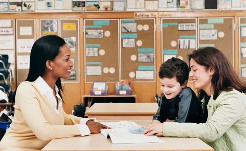 A teacher sitting on one side of the table showing a parent and student something in a textbook that are sat on the other side of the table. 