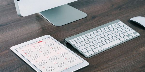 A tablet displaying a calendar next to a keyboard on a wooden desk.