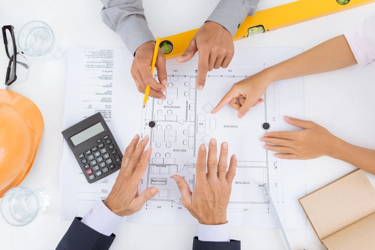 Team members meet to discuss company plans with a blueprint, calculator and hard hat on the table.