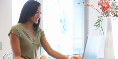 A woman working on a laptop in a bright room with a vase of flowers.