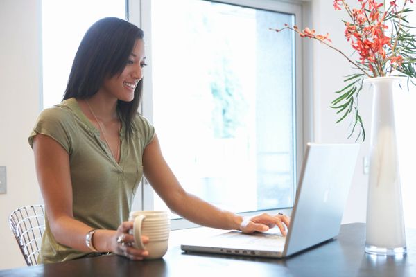 Woman working on laptop while holding a coffee mug at home.