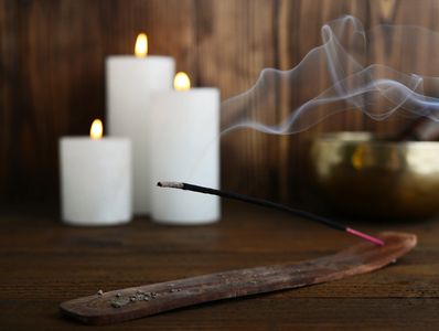 Lit incense stick with smoke and candles in the background on a wooden surface.