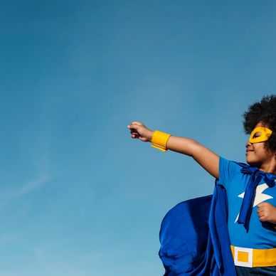 Child dressed as a superhero with a blue cape and mask against a clear sky.