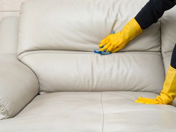Person cleaning a beige leather sofa with yellow gloves.