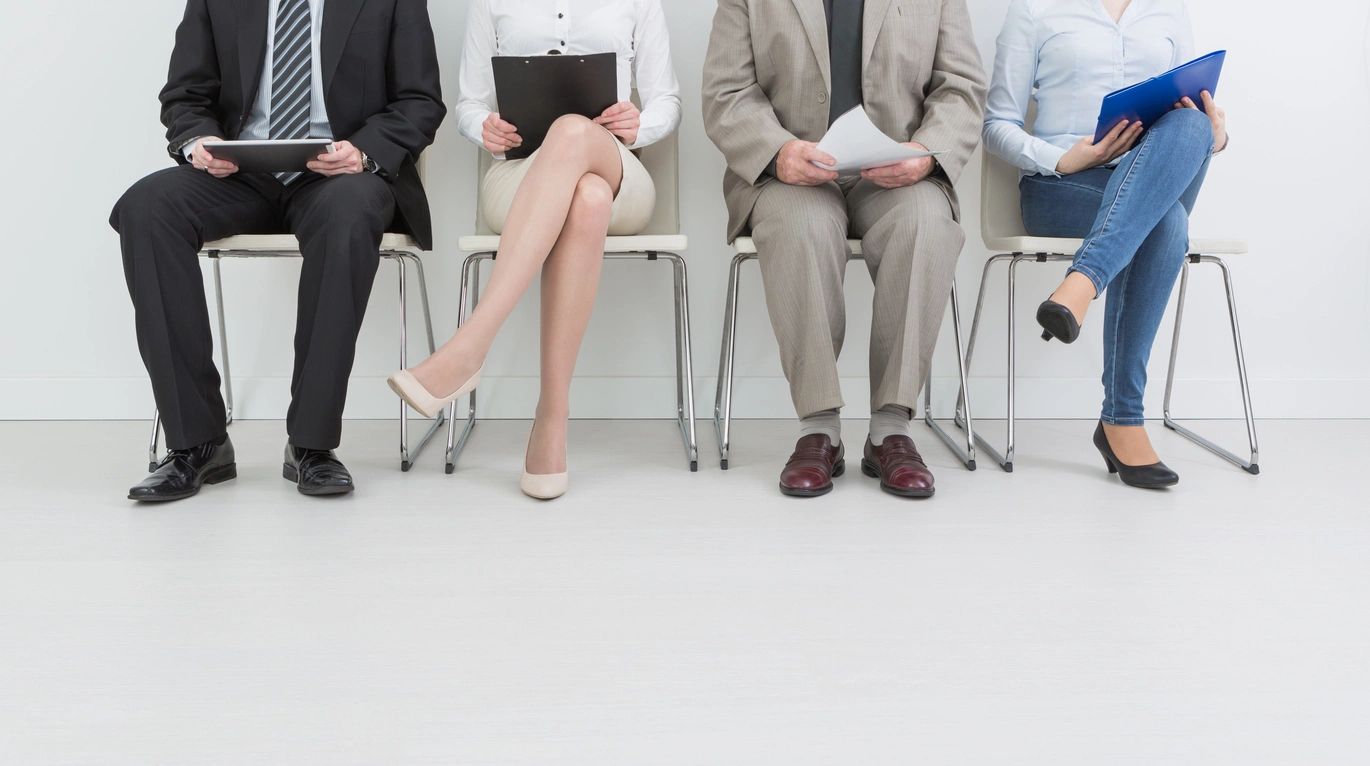 Four people sitting in chairs, waiting, holding documents.