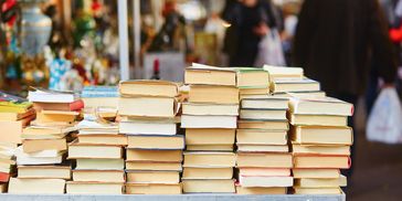 Stacks of used books on a market table with people in the background.