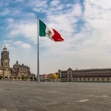 Mexican flag waving in a historic city square under a partly cloudy sky.