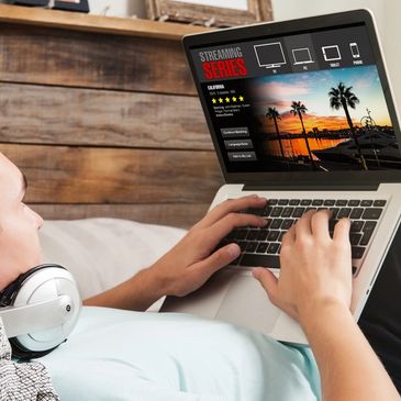 A person lying on a bed using a laptop with headphones around their neck.