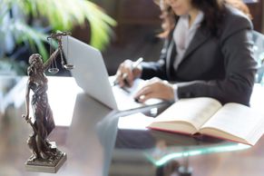 Woman at desk, laptop, open book, desk statue of Lady Justice.