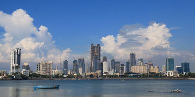 City skyline with a river in the foreground and scattered clouds in a blue sky.
