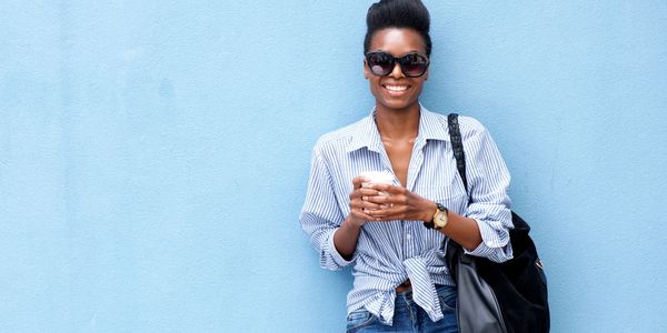 Smiling woman in sunglasses holding a coffee cup against a blue wall.