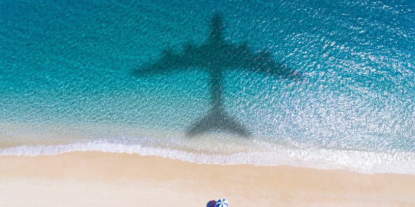 Shadow of an airplane over a beach with blue water and a person with an umbrella.