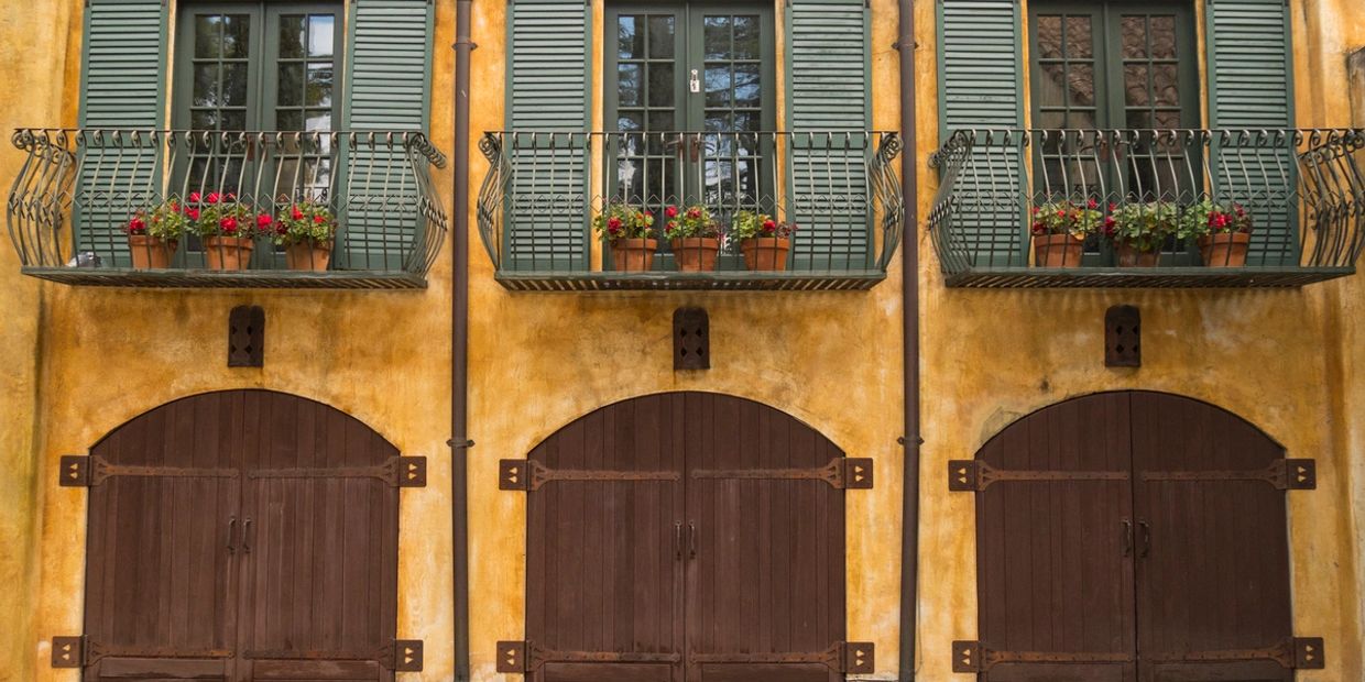Three balconies with flower pots and green shutters above arched wooden doors on a yellow building.