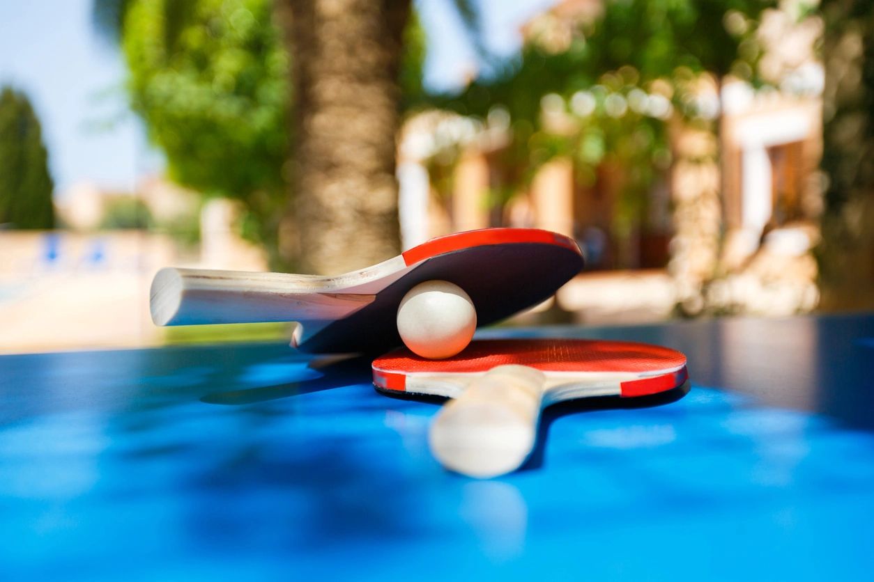 Two red ping pong paddles and a white ball on a blue table outdoors.