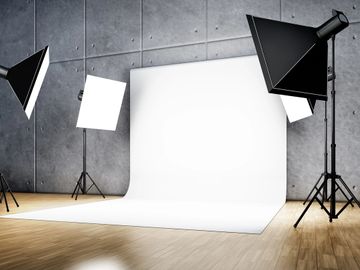 A professional photography studio setup with a seamless white backdrop, two softbox lights on stands to the left, and one softbox light on a stand to the right. The studio has a grey concrete wall and a wooden floor.