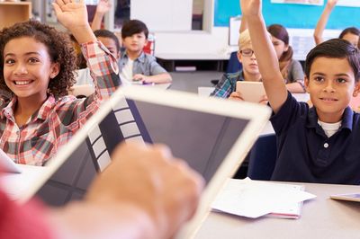 Students raising hands in a classroom with a tablet in the foreground.