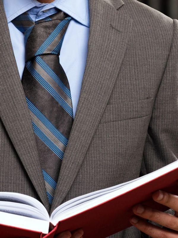 Man in a pinstripe suit and blue tie reading a red book.