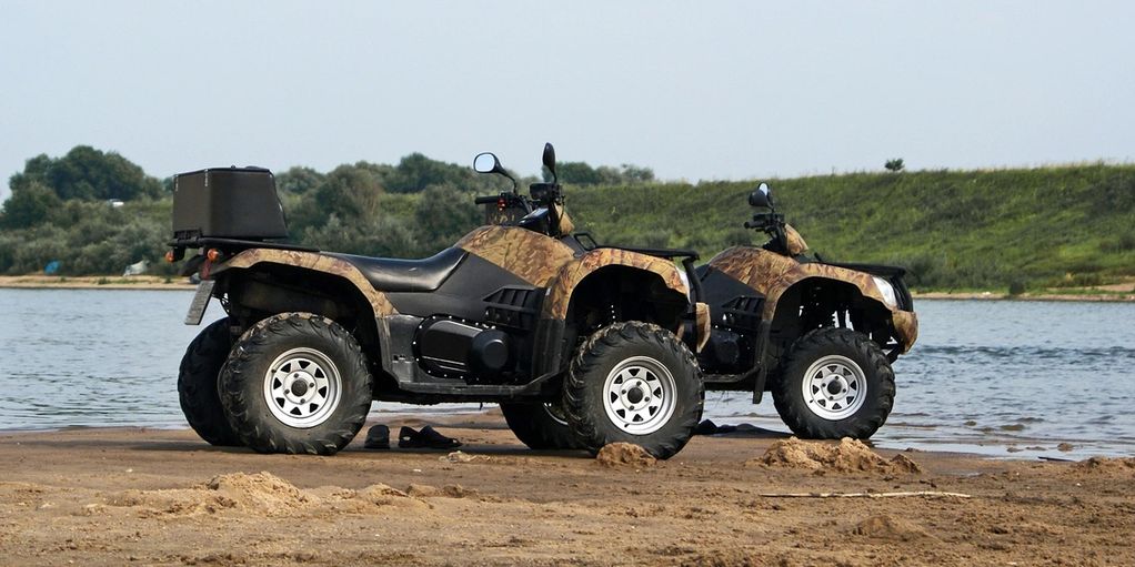 Two camouflaged all-terrain vehicles parked by a lakeside on sandy ground.