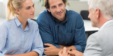 A man and woman on one side of a table speaking with a man on the opposite side. 