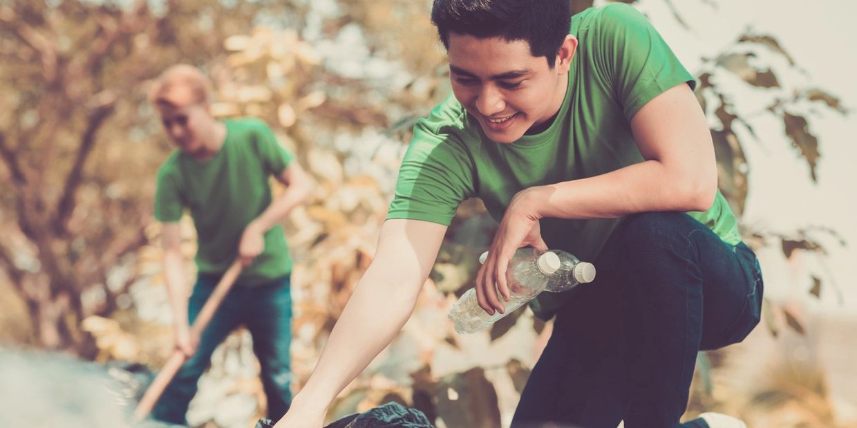 Two young men in green shirts picking up trash outdoors on a sunny day.