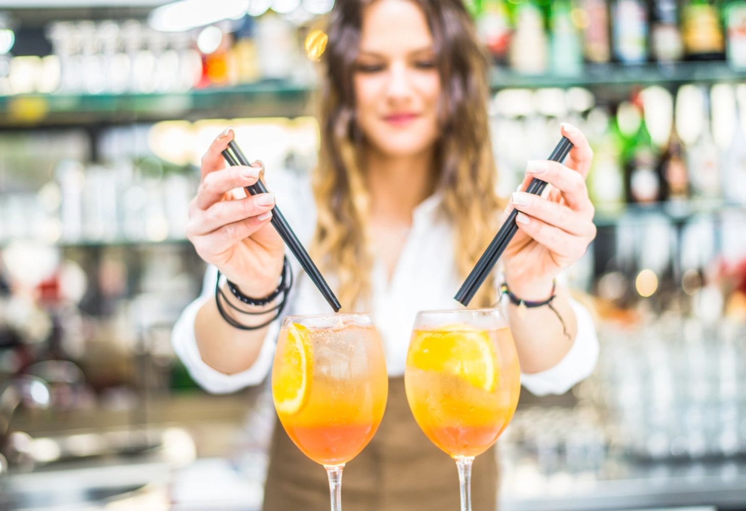 Bartender preparing two colorful cocktails with orange slices and straws.