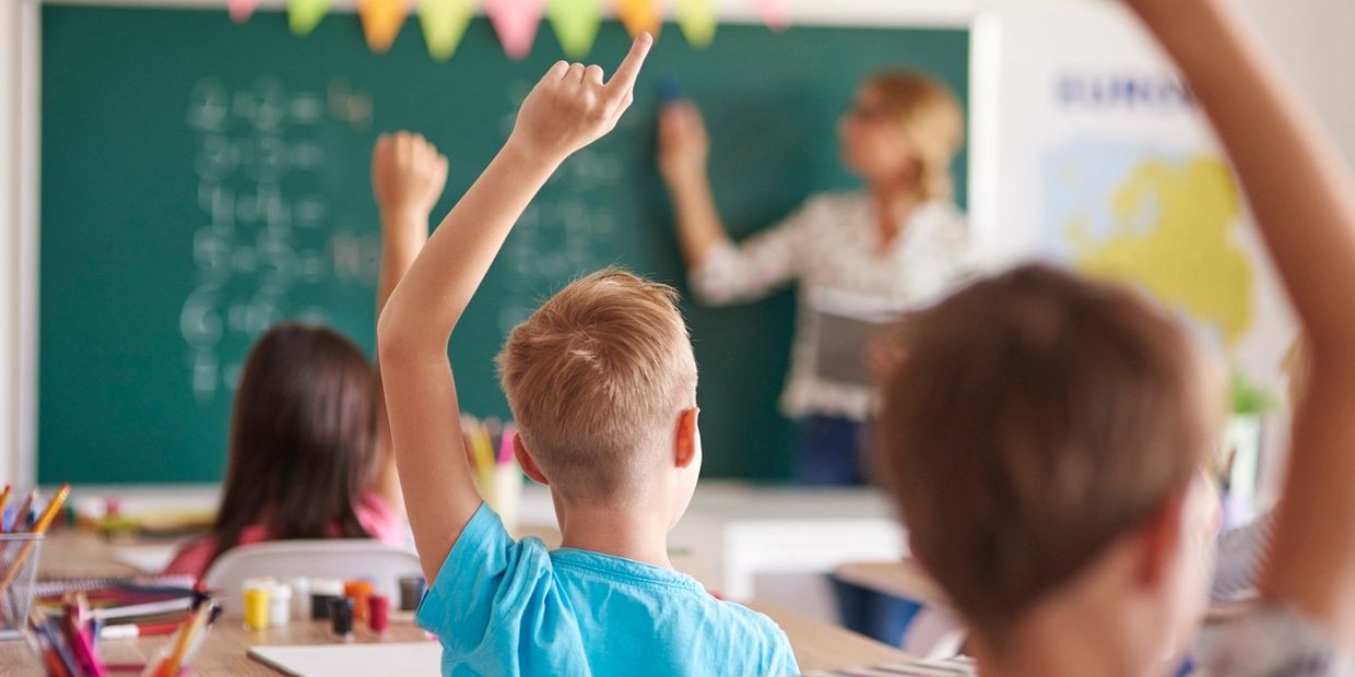 Students eagerly raise hands in classroom while teacher writes on chalkboard.