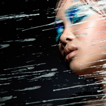 Close-up portrait of a woman with vibrant blue eye makeup behind streaked glass.