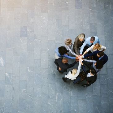 Eight people standing in a close circle, each holding an arm out to the centre and touching hands