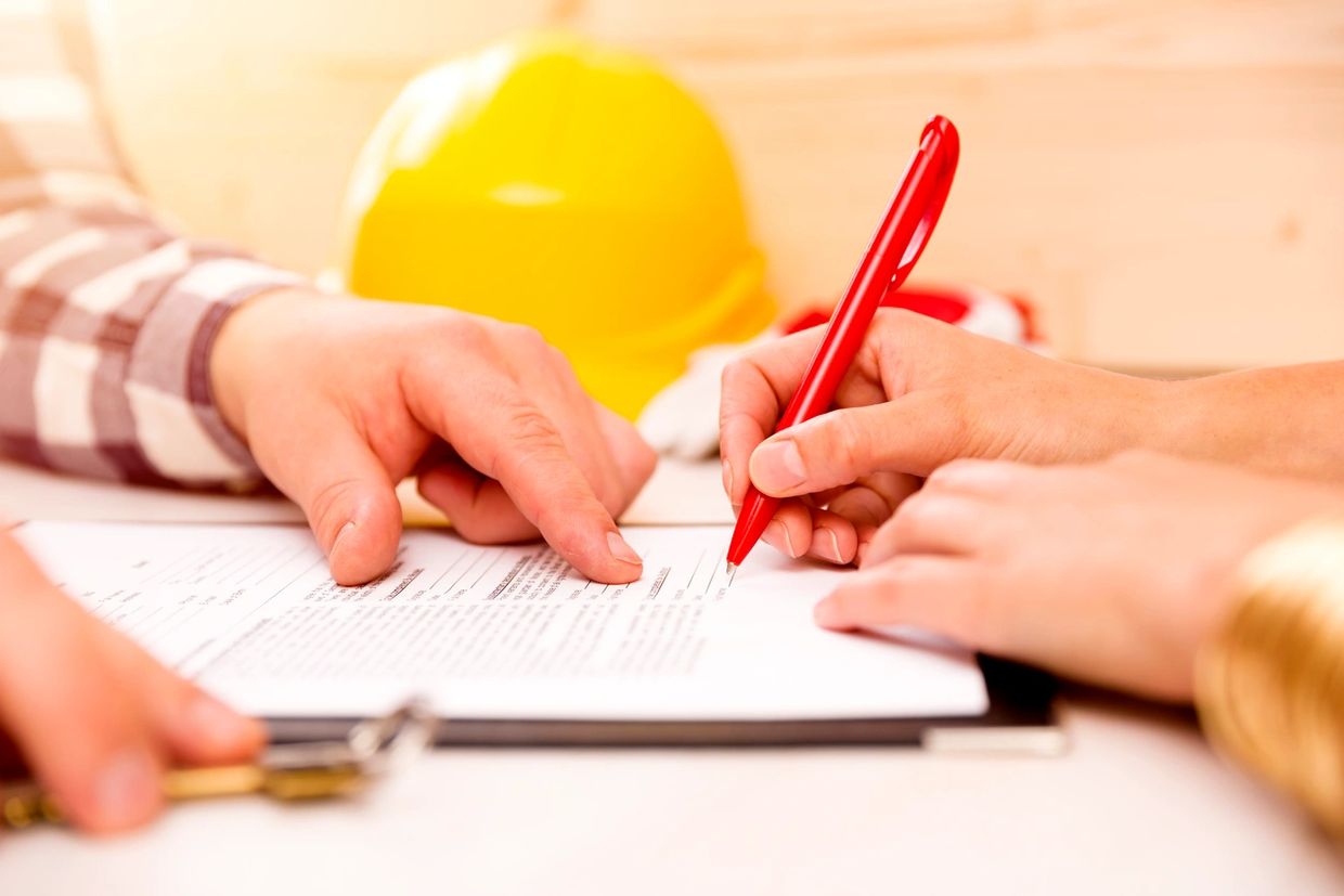 Hands signing a document with a red pen, construction helmet in background.