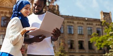 Two people using a laptop outdoors, smiling.