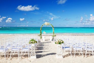 Beach wedding setup with white chairs and floral arch under a blue sky.
