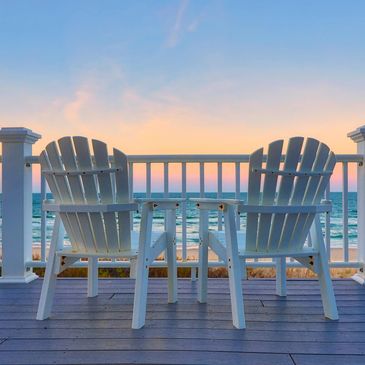 Two white chairs and a table on a deck overlooking the ocean at sunset.