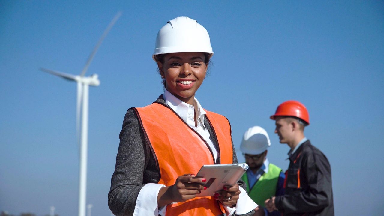 Technician stands in a wind turbine field with her hardhat on, tablet in-hand and two other co-workers.