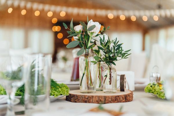 Elegant floral centerpiece with white roses and greenery in glass bottles on wooden slab.