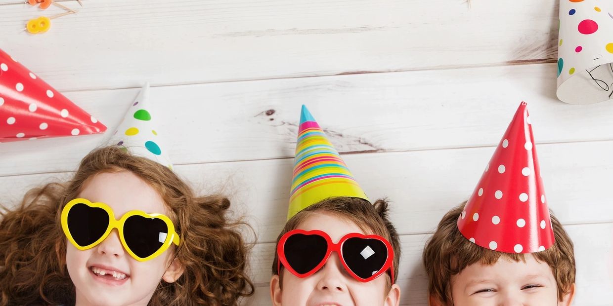 Three kids wearing colorful party hats and heart-shaped sunglasses, smiling happily.