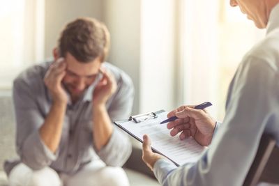 Man sitting leaning forward to fingers on side of face and man holding clipboard