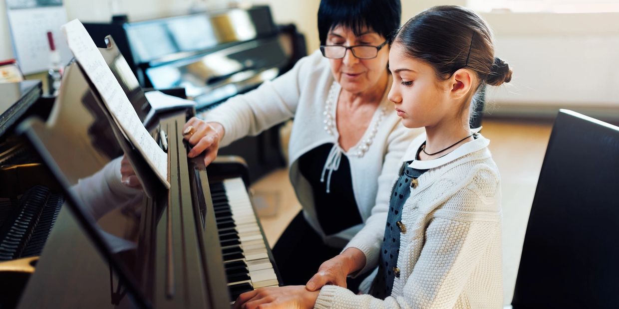 A woman teaching a young girl to play piano with sheet music in front.