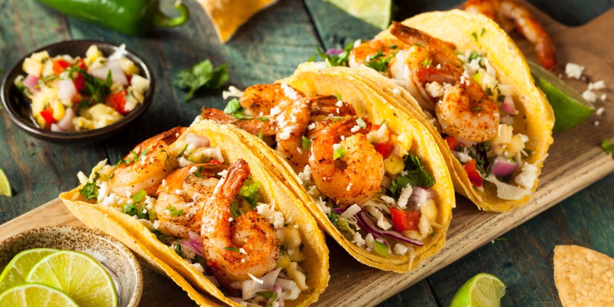 Three shrimp tacos with fresh salsa and lime wedges on a wooden board.