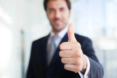 Smiling man wearing a black suit is blurry in the background.The image of him giving a big thumbs up