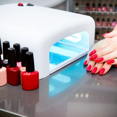 Hands with red nail polish drying under UV lamp next to nail polish bottles.