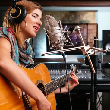 This is a image of a woman recording a song on a studio microphone while playing an acoustic guitar.