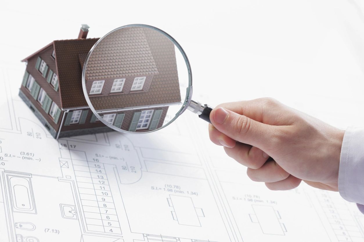 A magnifying glass is placed above a figurine of a home with a blueprint beneath it.