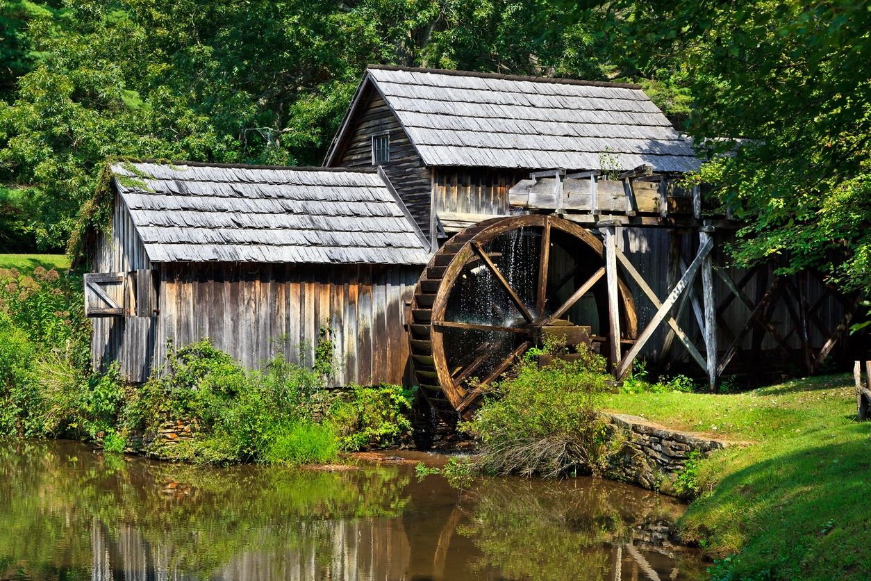 A rustic watermill by a pond surrounded by greenery.